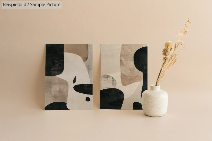 Two abstract paintings with beige and black patterns next to a white vase with dry flowers against a neutral background.