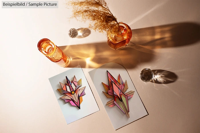 Two geometric floral designs on paper with amber glass vases casting shadows on a cream surface.