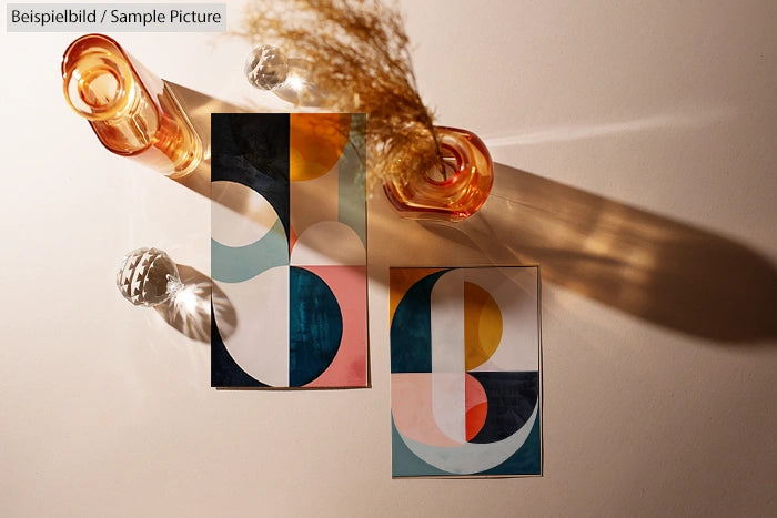 Two abstract geometric art prints with colorful shapes, next to amber glass vases casting shadows on a light surface.