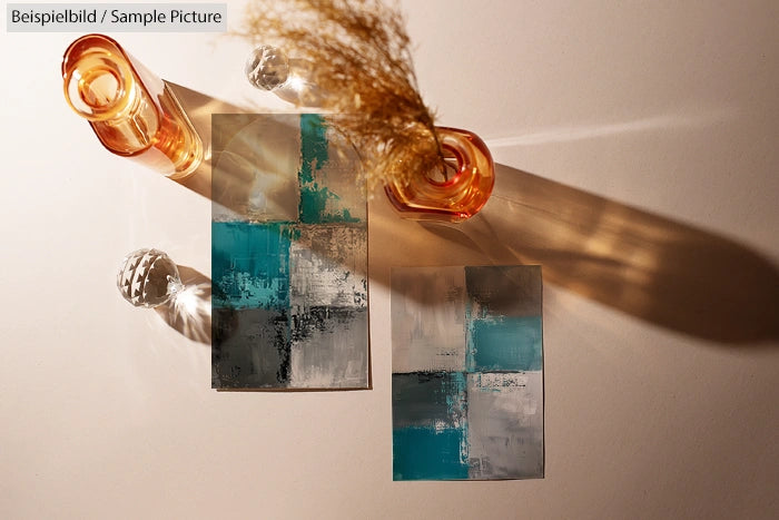 Abstract paintings with teal, gray, and white blocks next to amber glass vases casting shadows on a white surface.