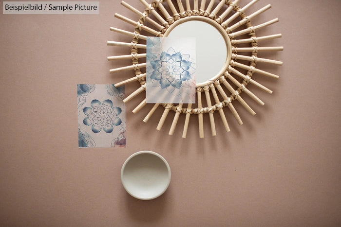 Round wicker mirror on pink surface with floral sketches and small white bowl.