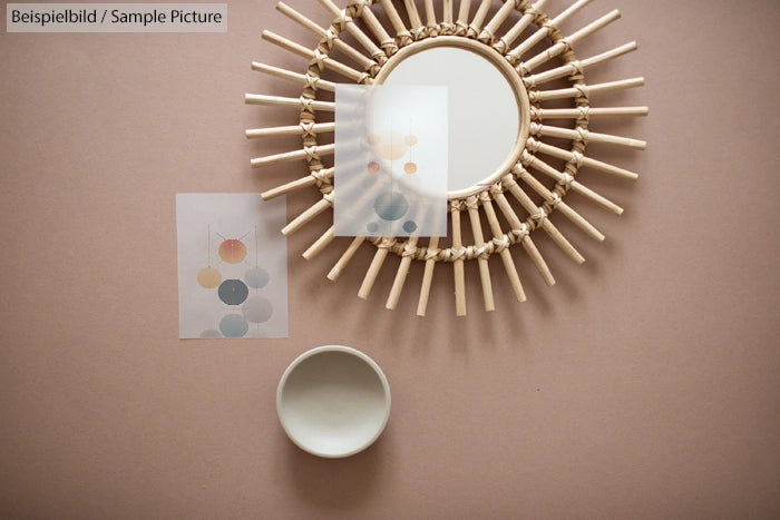 Round mirror with wicker frame, abstract art cards, and a small white bowl on a pinkish surface.