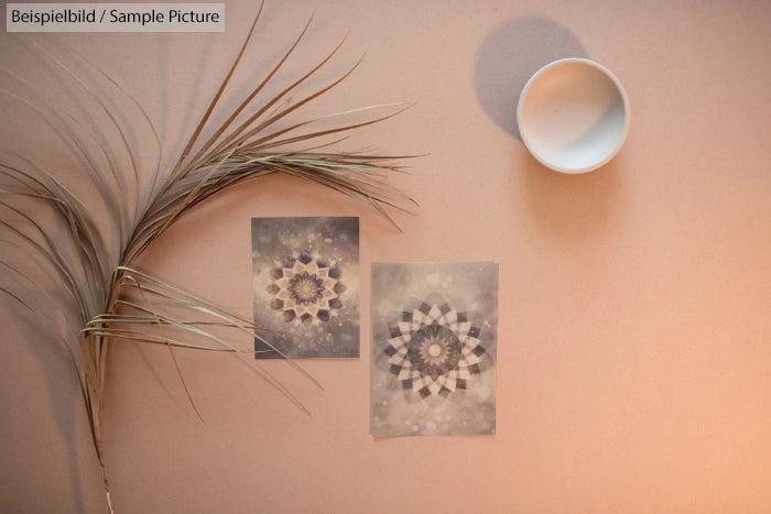 Two abstract mandala prints on a beige surface with a dry plant and a white bowl nearby.