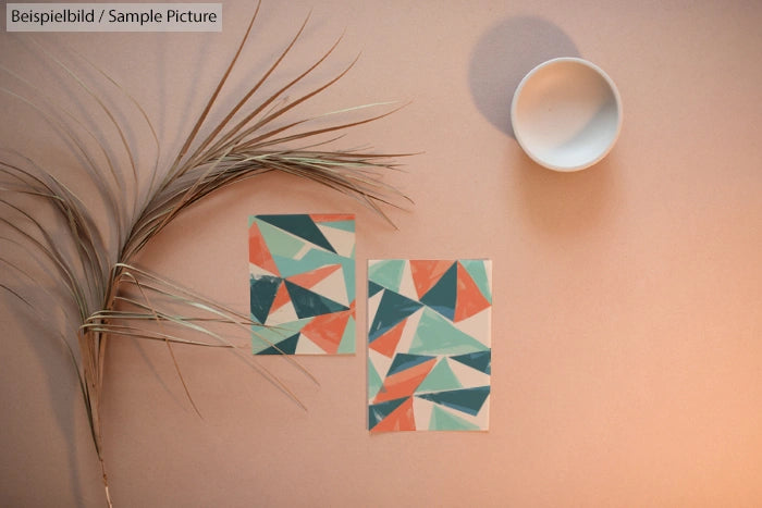 Geometric abstract art prints with teal and coral triangles on beige background, accompanied by dried plant and white bowl.