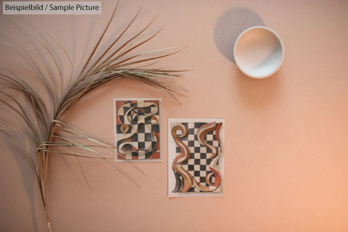 Decorative abstract checkered art prints on beige background with dried plant and round white object.