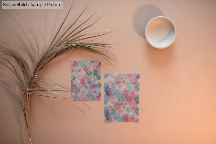 Two patterned papers on table with dried plant and empty cup, soft lighting.