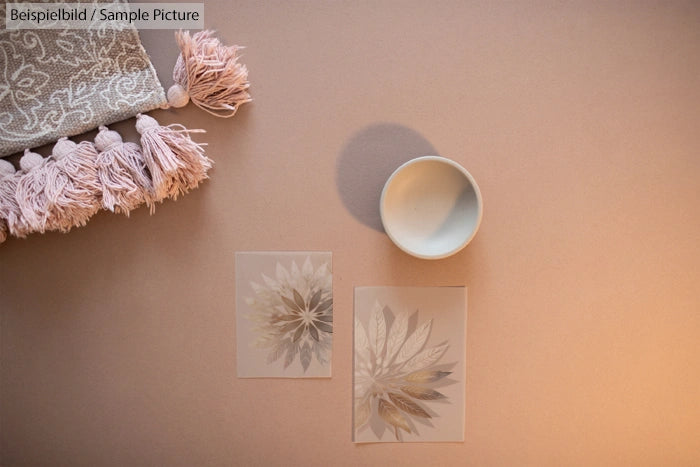 Decorative tassel cloth with a ceramic bowl and two transparent floral sheets on a beige surface.