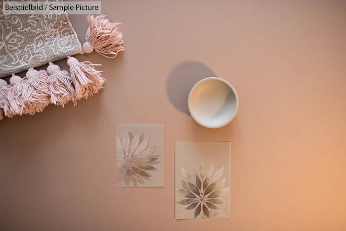 Decorative tassels with two floral design sheets and a white ceramic cup on a peach background.