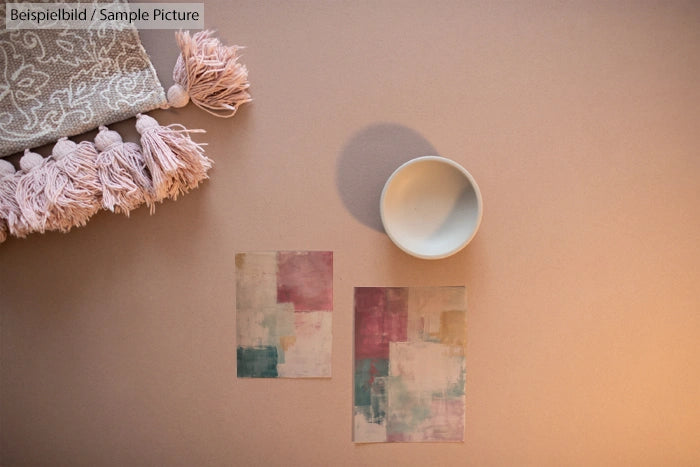 Aerial view of a ceramic bowl on paper with abstract pastel art and a tasseled cloth on a neutral background.