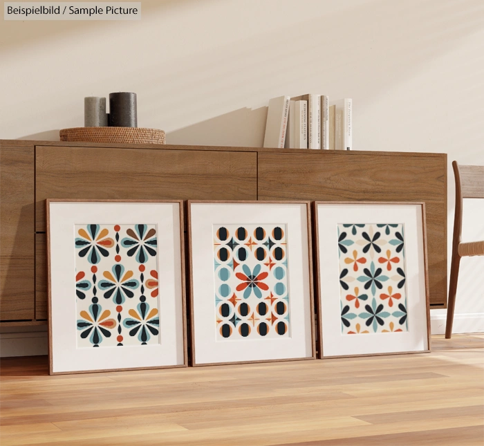 Three framed geometric prints on a wooden floor by a modern sideboard with books and decor items.