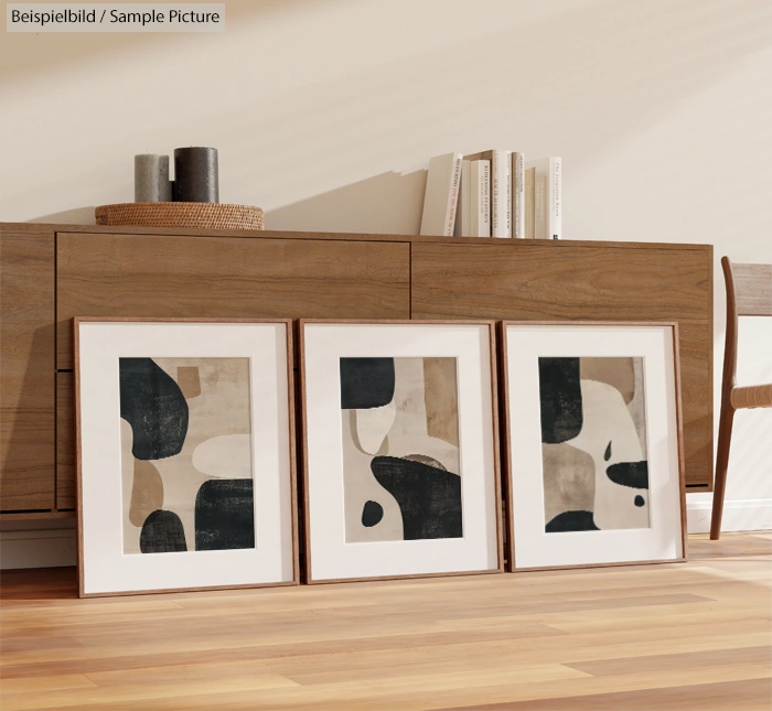 Three framed abstract artworks on a wooden floor in front of a wooden sideboard.