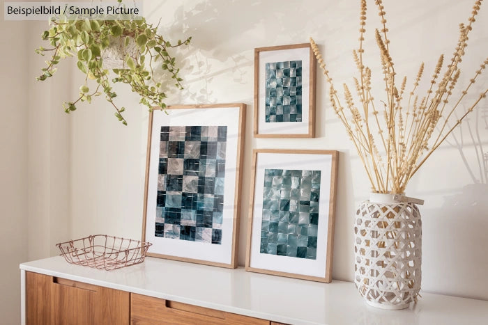 Three framed abstract artworks on a sideboard with a plant and decorative vase.