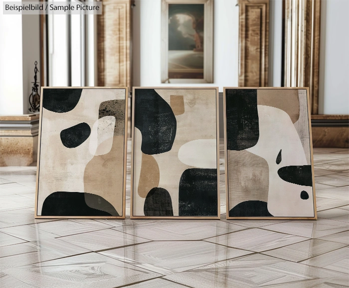 Three abstract paintings with black, cream, and beige shapes in wooden frames on a polished floor.