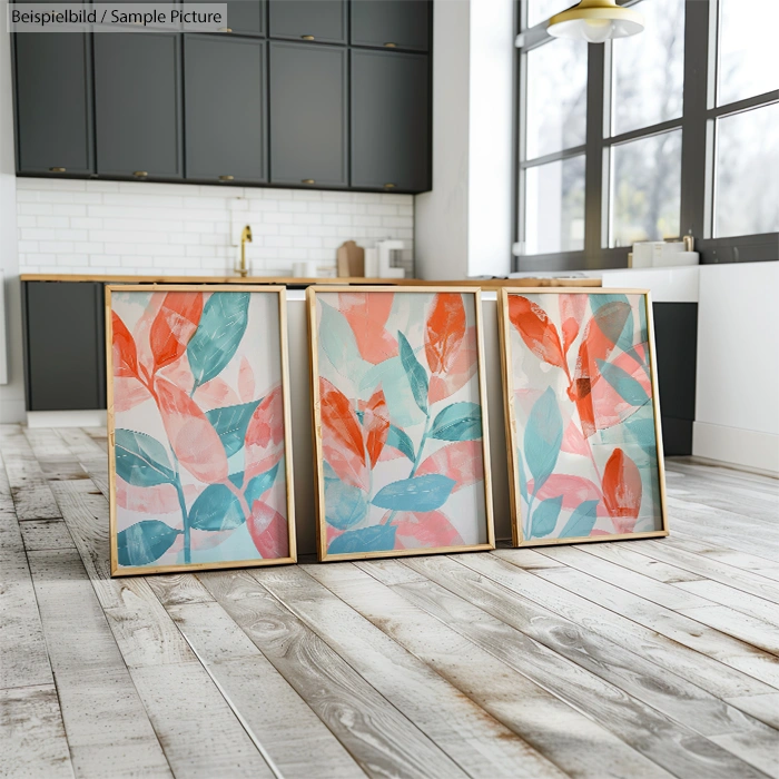 Three abstract leaf paintings in vibrant blue, pink, and orange hues displayed on a wooden floor in a modern kitchen.