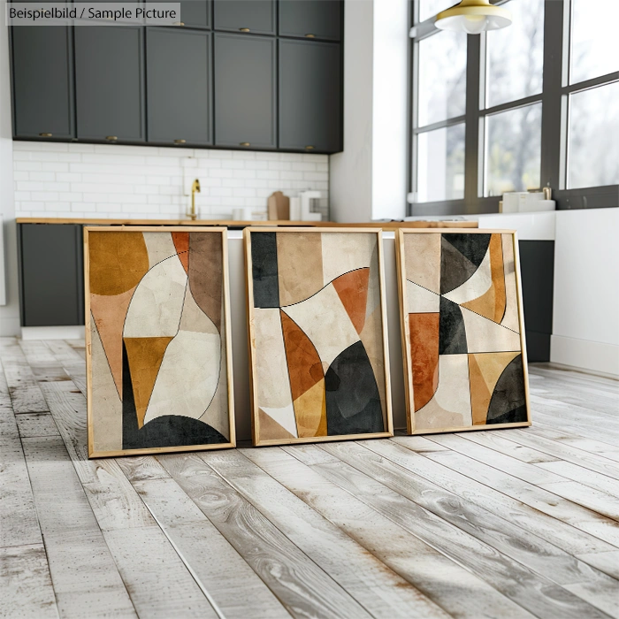 Three framed abstract paintings with geometric shapes in earth tones on a wooden floor in a modern kitchen.