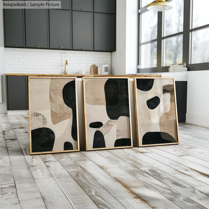 Three abstract paintings with beige, black, and brown shapes leaning against a kitchen counter in a modern room.