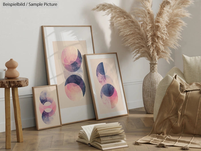 Framed abstract artworks with geometric patterns, placed on wooden floor beside pampas grass and cushions.