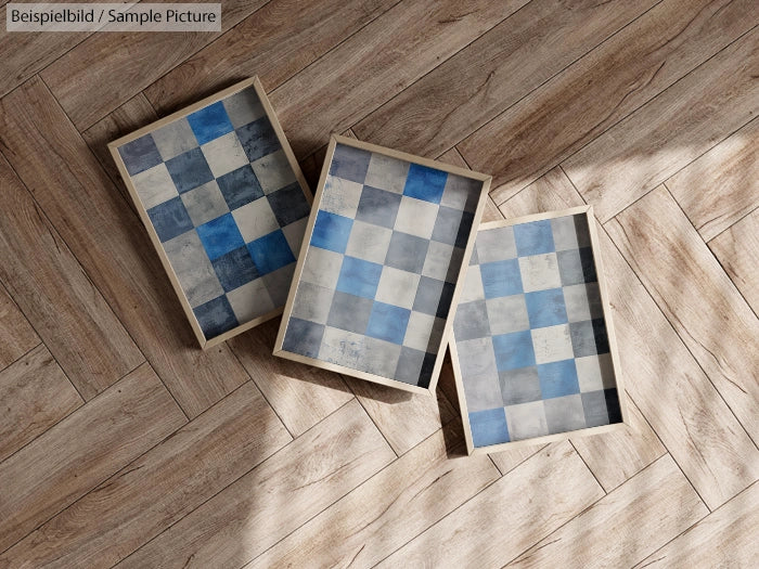 Three framed blue and gray checkered prints on a wooden floor with diagonal sunlight.