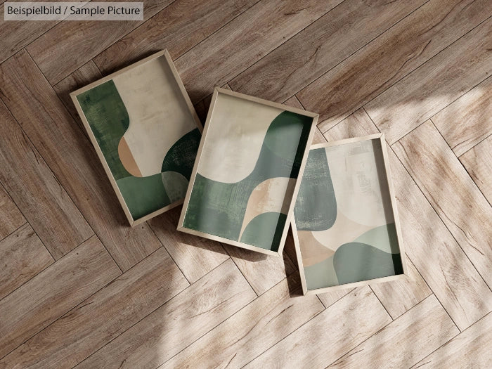 Three framed abstract paintings with green and beige curves placed on a wooden floor.