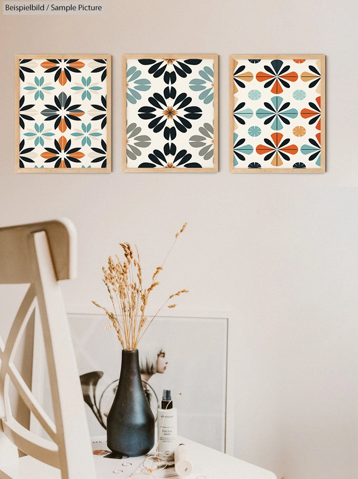 Wall with three framed geometric art pieces above a white table with a dark vase and dried flowers.