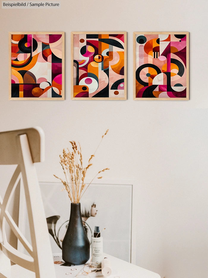 Modern art prints with geometric shapes in warm tones on a wall above a table holding vase with dried flowers.