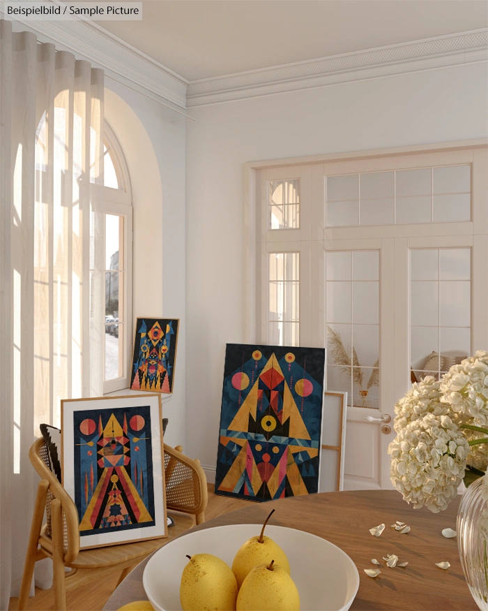 Modern art paintings on display in a bright room with white flowers and pears on a table, beside large windows.