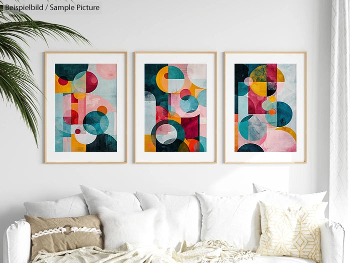 Three framed abstract art prints with geometric shapes in a white room above a white couch with beige and white pillows.