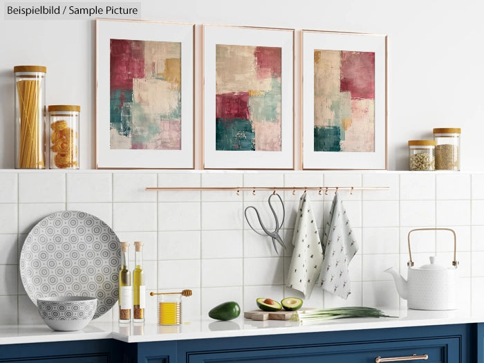 Kitchen countertop with blue cabinets, abstract art, white kettle, plates, avocados, and hanging utensils.