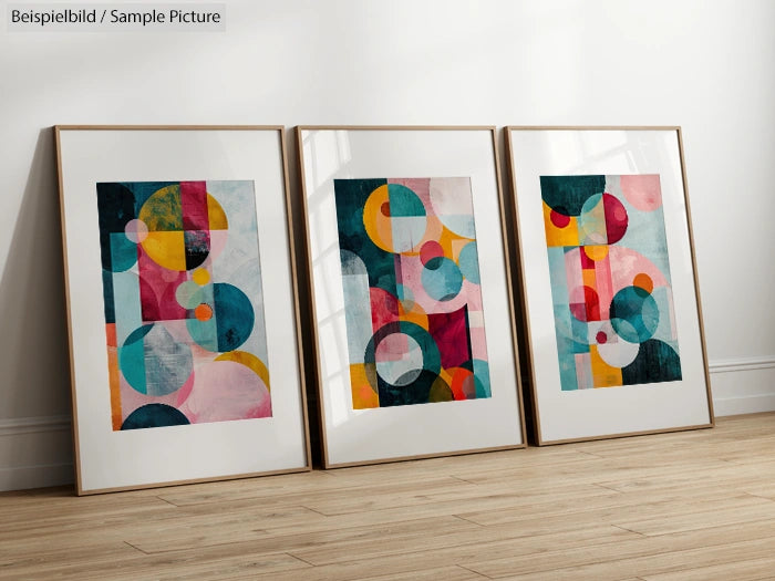 Three framed abstract art pieces with geometric shapes in vibrant colors leaning against a wall on a wooden floor.